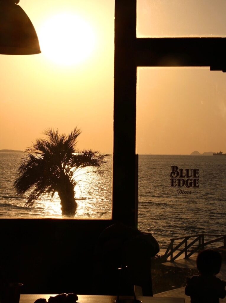 BLUE EDGE Diner.の店内窓から見える夕暮れの日本海とヤシの木のシルエット