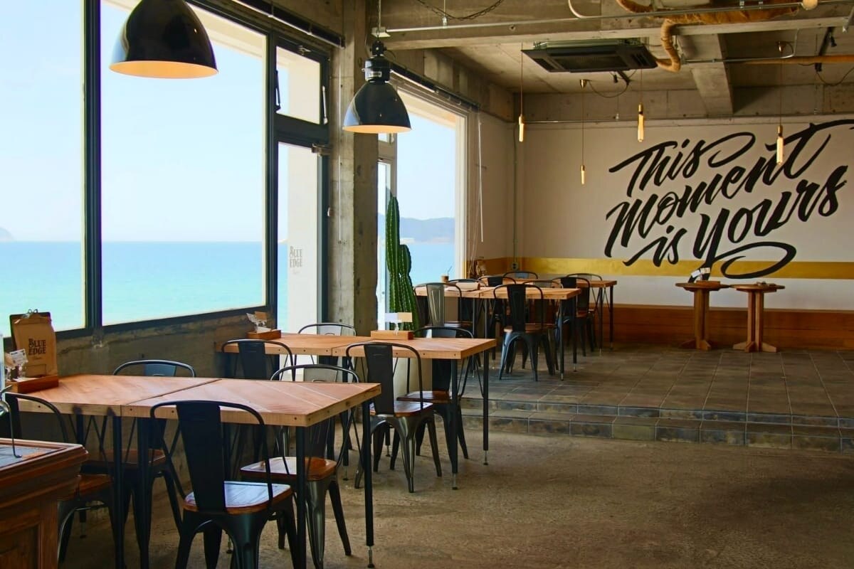 BLUE EDGE Diner.の店内、海側の大きな窓と木製テーブル席、壁のロゴアート(公式サイトより)