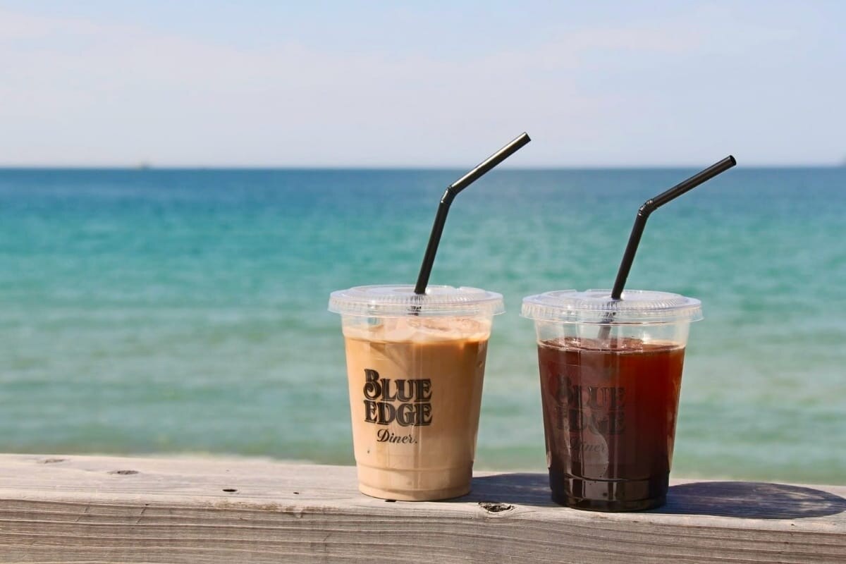 BLUE EDGE Dinerのアイスコーヒーとアイスティー、海辺の手すりに置かれたテイクアウトカップ(公式サイトより)