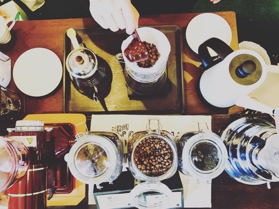 CAFE麓のコーヒーカウンター。ドリッパーに豆を入れる手元と、ケトル、コーヒーミル、コーヒー豆の入ったガラス瓶が並ぶ(公式サイトより)