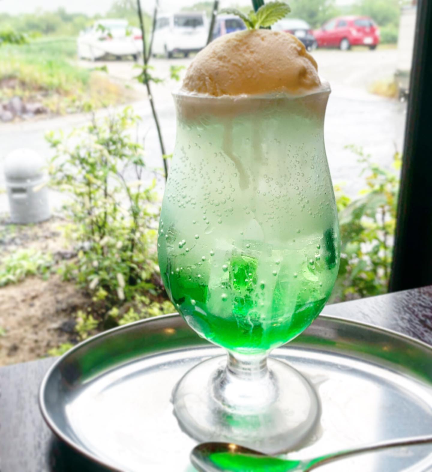 CAFE麓のメロンクリームソーダ。緑から白へのグラデーションのソーダにアイスとミントがのり、窓の外に緑が見える(公式Instagramより)