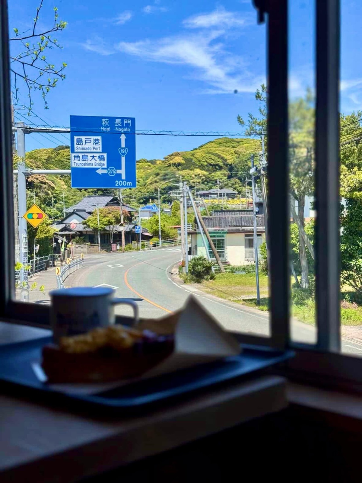窓辺のトレーにマグカップとホットドッグ。窓の外には青空と緑の山、「角島大橋 275 200m」「島戸港」「萩 長門 191」と書かれた青い道路標識が見える（Goodies Ella公式Instagramより）