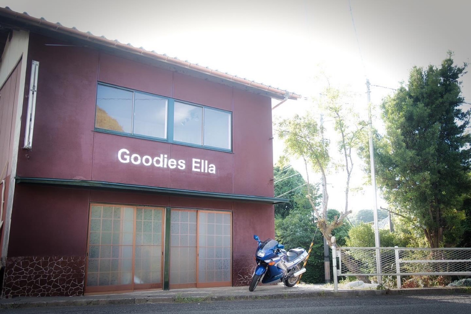 「Goodies Ella」の白い文字が入ったえんじ色の2階建ての建物と、店先に停められたバイク（Goodies Ella公式Instagramより）