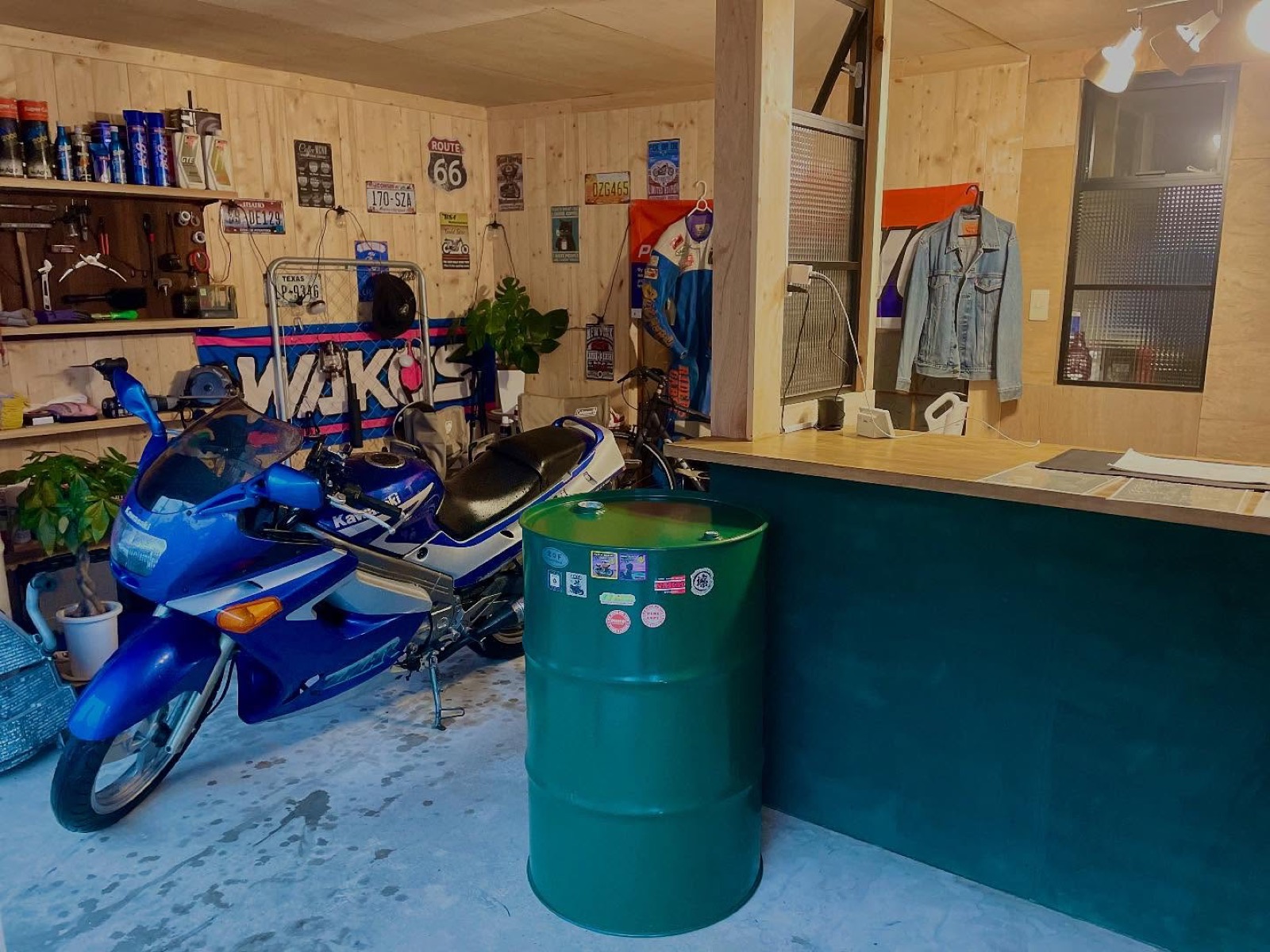 コンクリートの床に青いバイクと緑のドラム缶が置かれ、木の壁にジャケットやポスターが飾られたカウンターまわりの店内（Goodies Ella公式Instagramより）