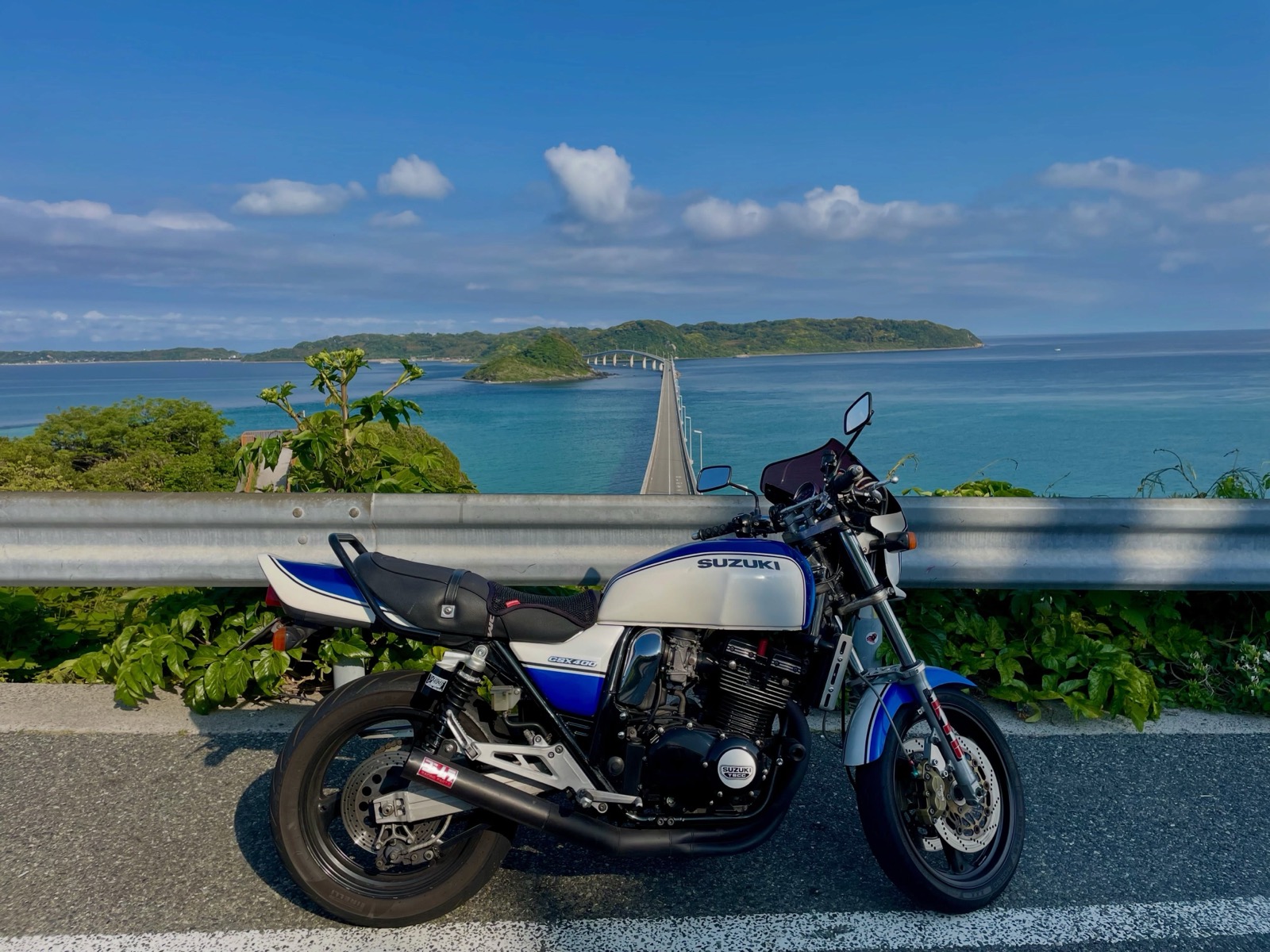 ガードレール沿いに停めた青と白のバイク。背景にはエメラルドブルーの海と、島へまっすぐ伸びる角島大橋が見える（Goodies Ella公式Instagramより）