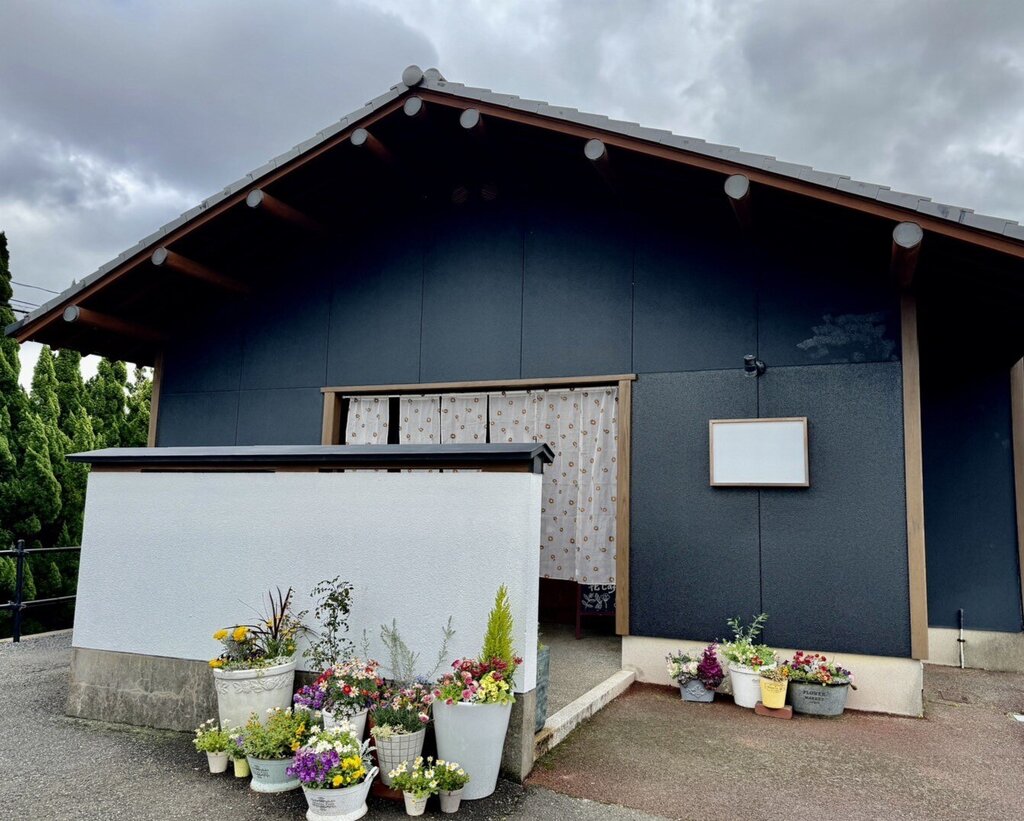 黒い切妻屋根の古民家風の建物「花cafe」の外観。白い塀の前に鉢植えの花々が並び、暖簾のかかった入り口が見える（花cafe公式サイトより）