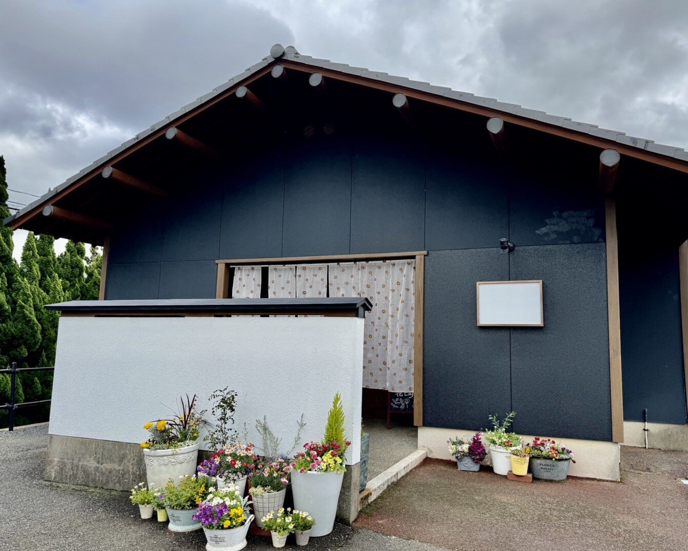黒い切妻屋根の古民家風の建物「花cafe」の外観。白い塀の前に鉢植えの花々が並び、暖簾のかかった入り口が見える(花cafe公式サイトより)