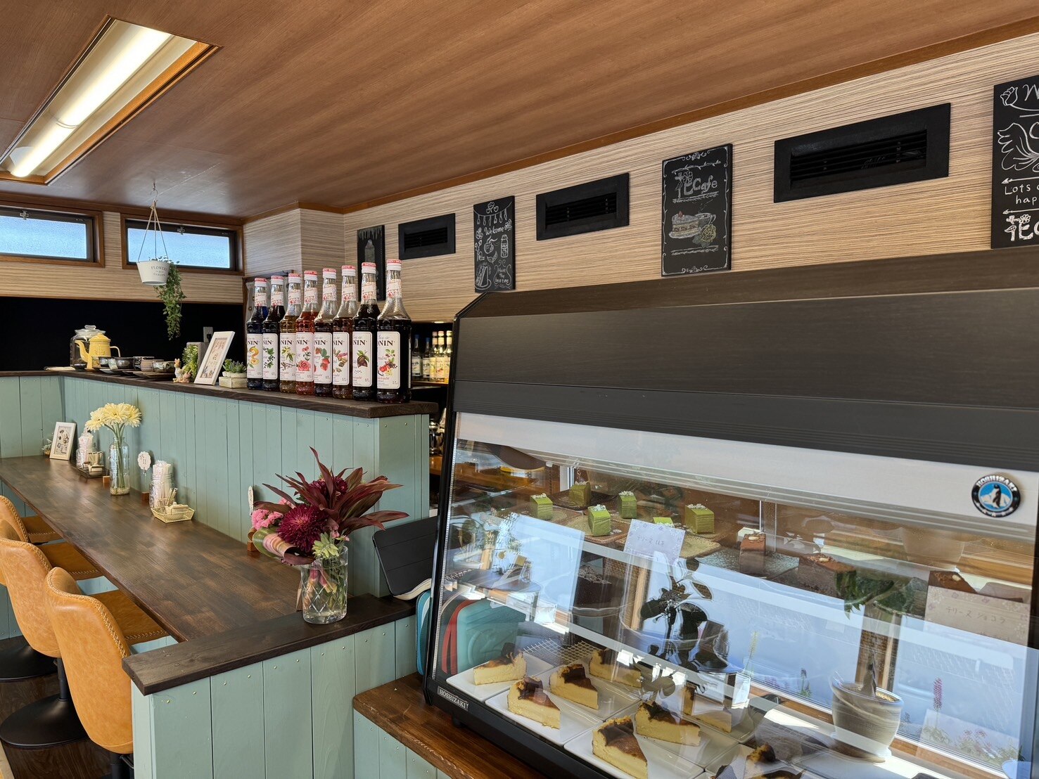 花cafeの店内。ミントグリーンのカウンターと木目の壁に囲まれたケーキケースに焼き菓子が並び、黄色い椅子が置かれている(花cafe公式サイトより)