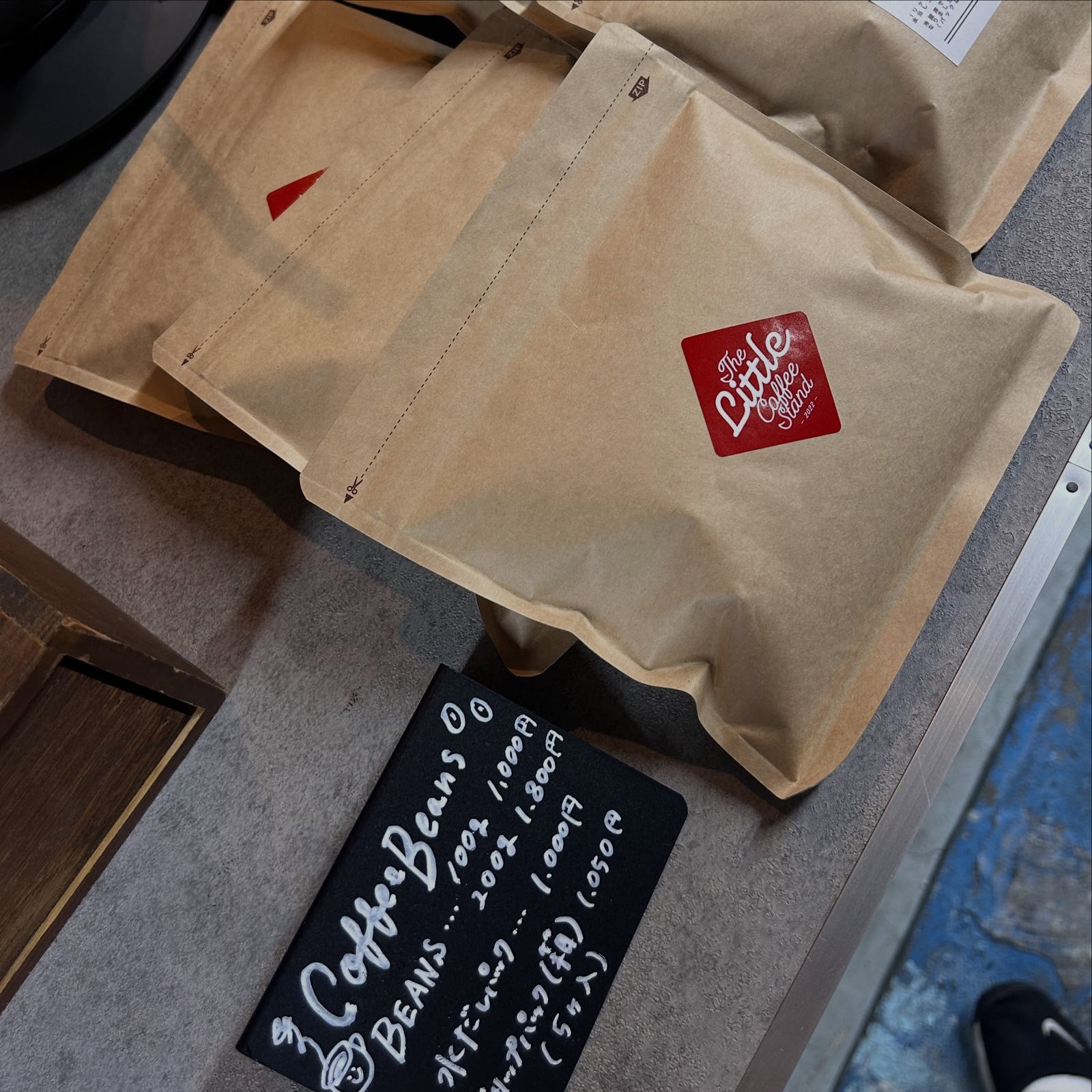 赤いロゴシールが貼られたクラフト袋のコーヒー豆と、Coffee Beansの価格が書かれた黒板(The Little Coffee Stand公式Instagramより)