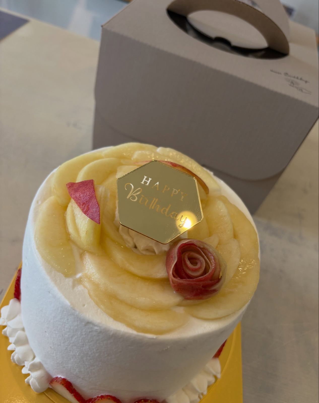 薄切りの桃を花びらのように重ね、赤いバラ形の飾りとHAPPY Birthdayのプレートをのせた白いクリームのシフォンデコレーションケーキ（Sana all公式Instagramより）