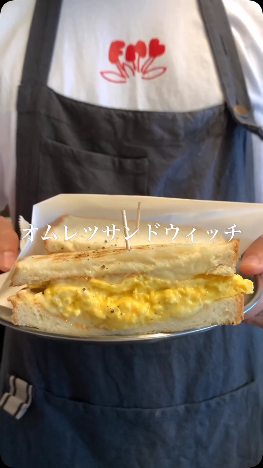 「オムレツサンドウィッチ」の文字入り。エプロン姿のスタッフが、黄色いふわふわの卵をはさんだトーストサンドをお皿にのせて持っている(Sh:AKARIKI COFFEE公式Instagramより)