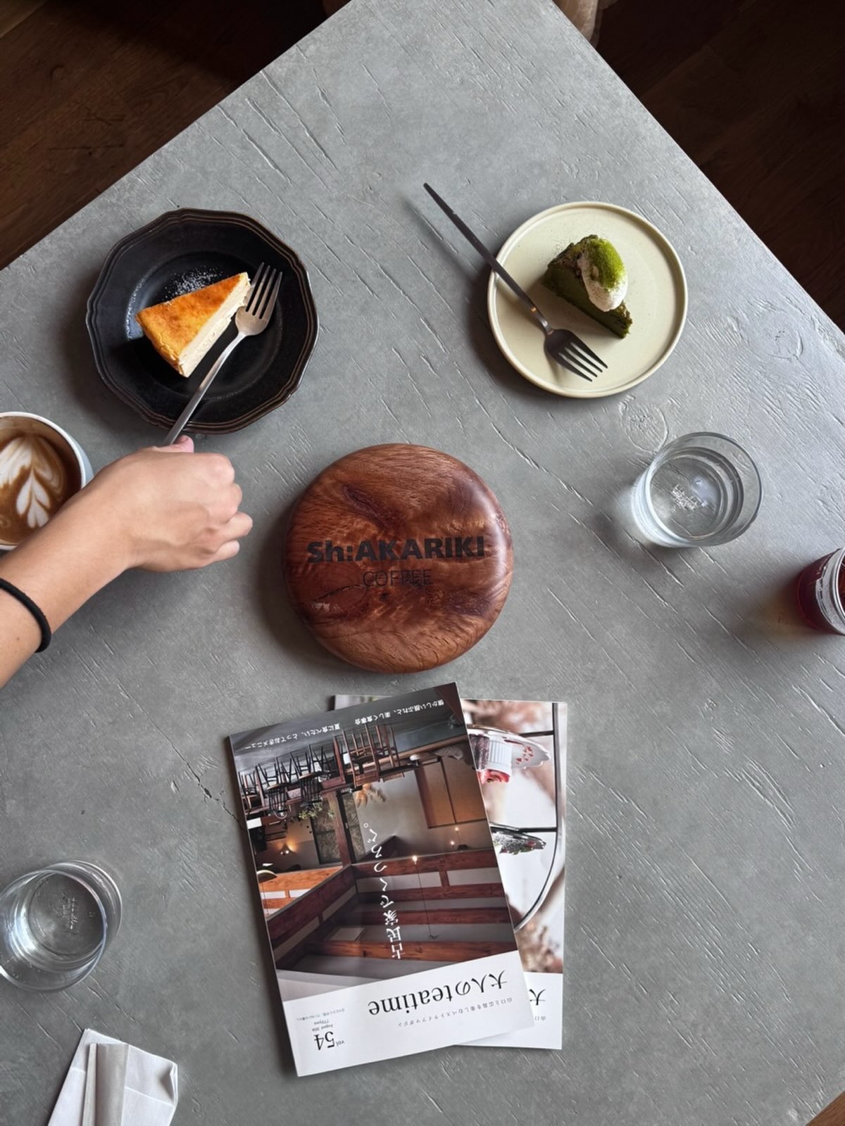 グレーのテーブルの上に、焼き色のついたケーキと抹茶色のケーキ、ラテアートのカップ、Sh:AKARIKI COFFEEのロゴ入りの木のコースター、雑誌が並ぶ様子を真上から撮った写真(Sh:AKARIKI COFFEE公式Instagramより)