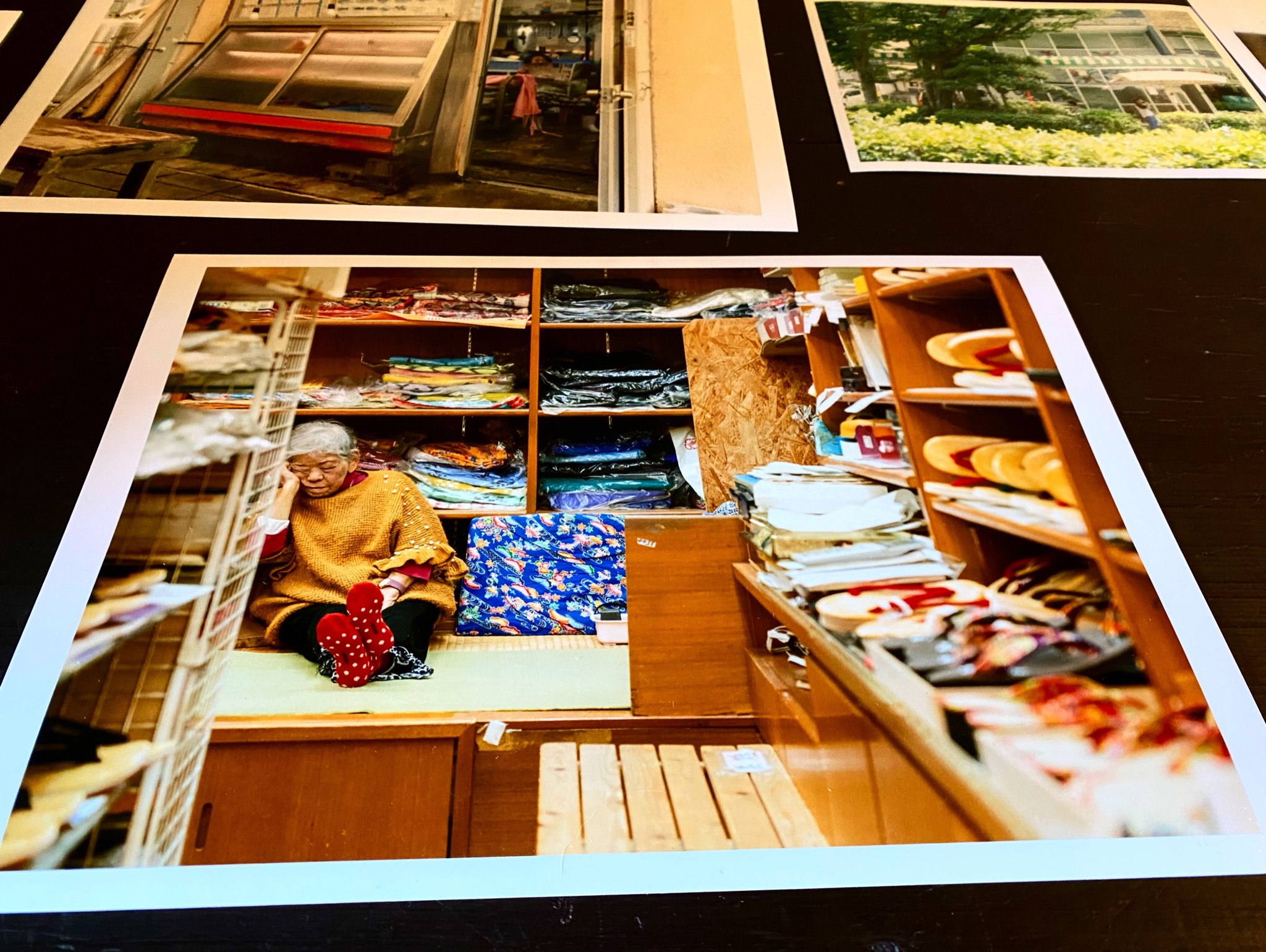 黒いテーブルの上に並べられた写真プリント。呉服店の店内らしき場所に座る女性を写した作品が中央に置かれている(Gallery CAFE とこのま公式Instagramより)