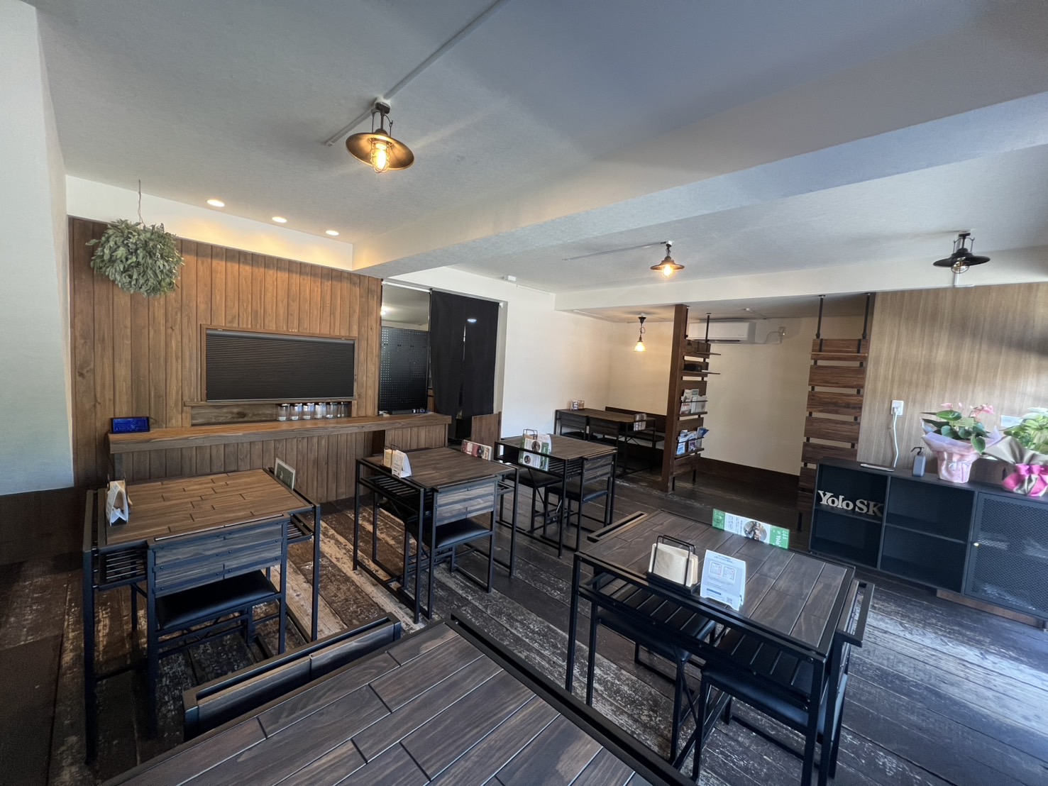 Yolo's cafeの店内。ダークブラウンの木の床にアイアンフレームの四角いテーブル席が並び、アンティーク調のペンダントライトが下がる(合同会社YoloSK公式サイトより)