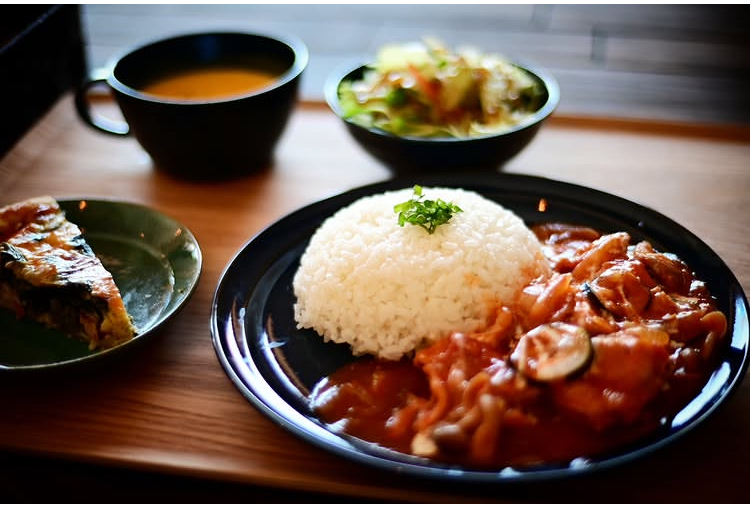 黒い平皿に盛られたトマト煮込みとライス、緑を散らしたごはん。奥にポタージュスープのカップとサラダの小鉢、左手前に緑の皿にのったキッシュ(合同会社YoloSK公式サイトより)