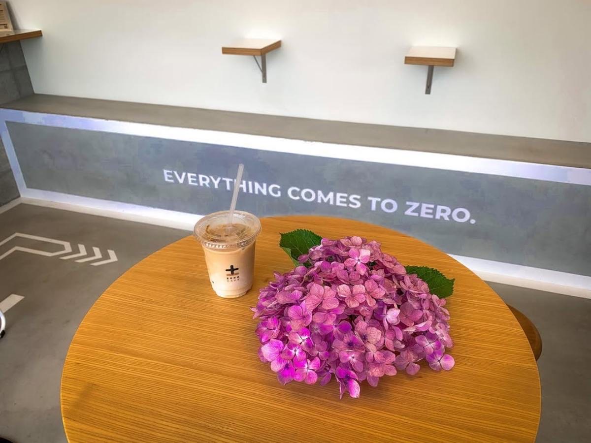 店内の木の丸テーブルに置かれたZERO CAFEロゴ入りカップのアイスラテと、ピンクのアジサイの花（ZERO CAFE公式サイトより）