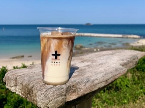 流木のテーブルに置かれたロゴ入りカップのアイスラテと、背景に広がる青い海（ZERO CAFE公式サイトより）