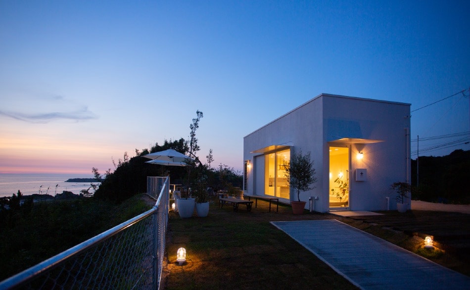 夕暮れの空の下、窓と入口に明かりが灯るZERO CAFEの建物と、足元のライトに照らされた芝生のテラス（ZERO CAFE公式サイトより）