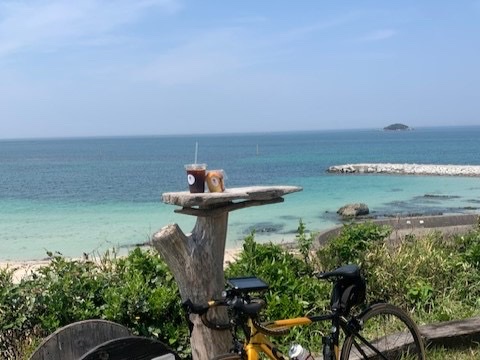 海を望む流木のテーブルに置かれたアイスコーヒーとドリンク、手前に停められた自転車（ZERO CAFE公式サイトより）