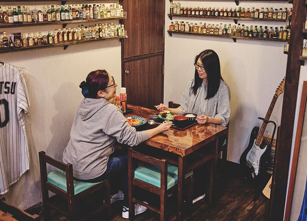 なすび食堂の店内。レコードやウイスキーボトルが並ぶレトロな内装でテーブル席につく2人の客(公式サイトより)