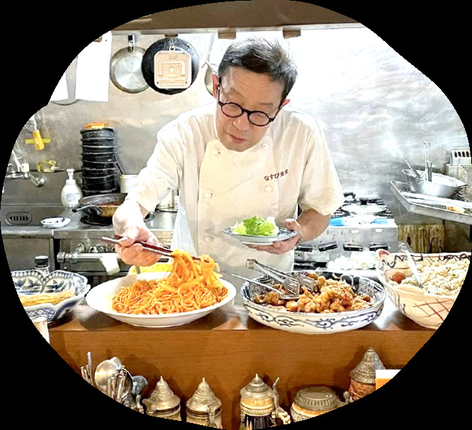 厨房でナポリタンやから揚げを盛り付けるなすび食堂の店主(公式サイトより)